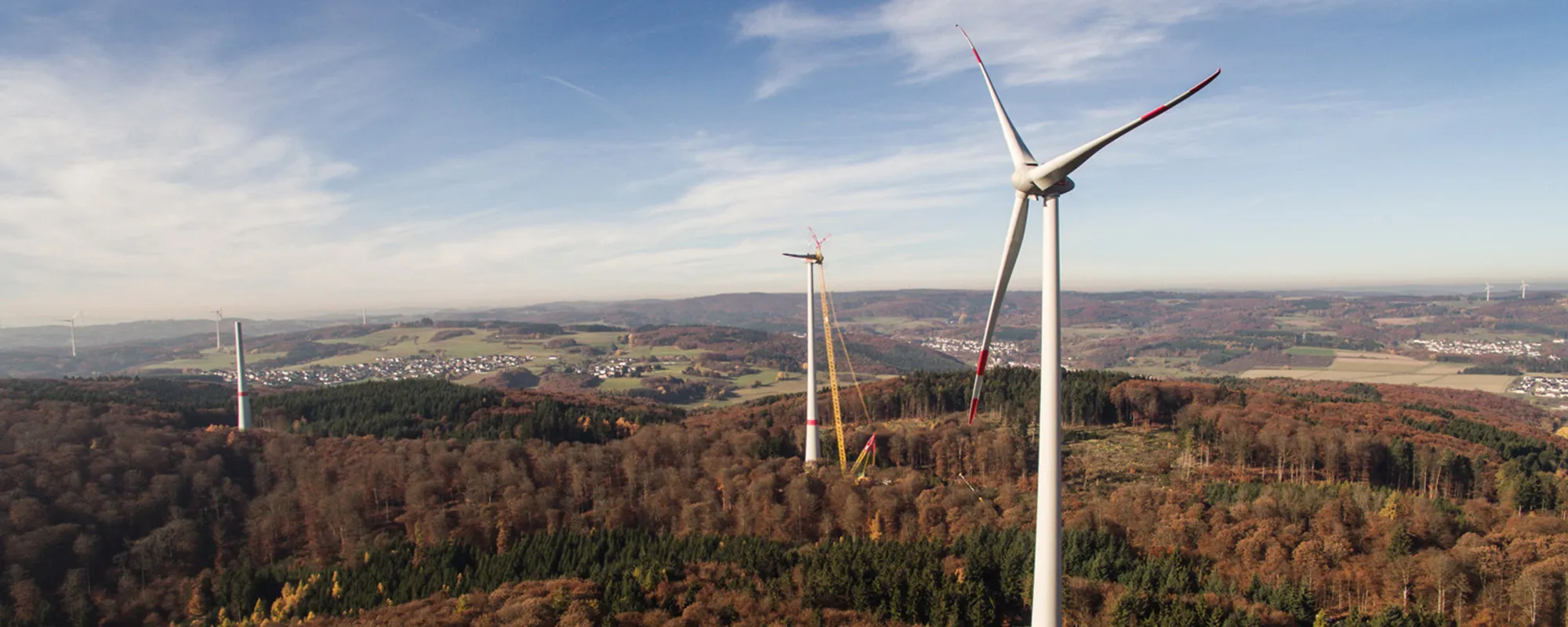 Luftaufnahme von drei Windkraftanlagen in einer herbstlichen Waldlandschaft mit sanften Hügeln im Hintergrund.