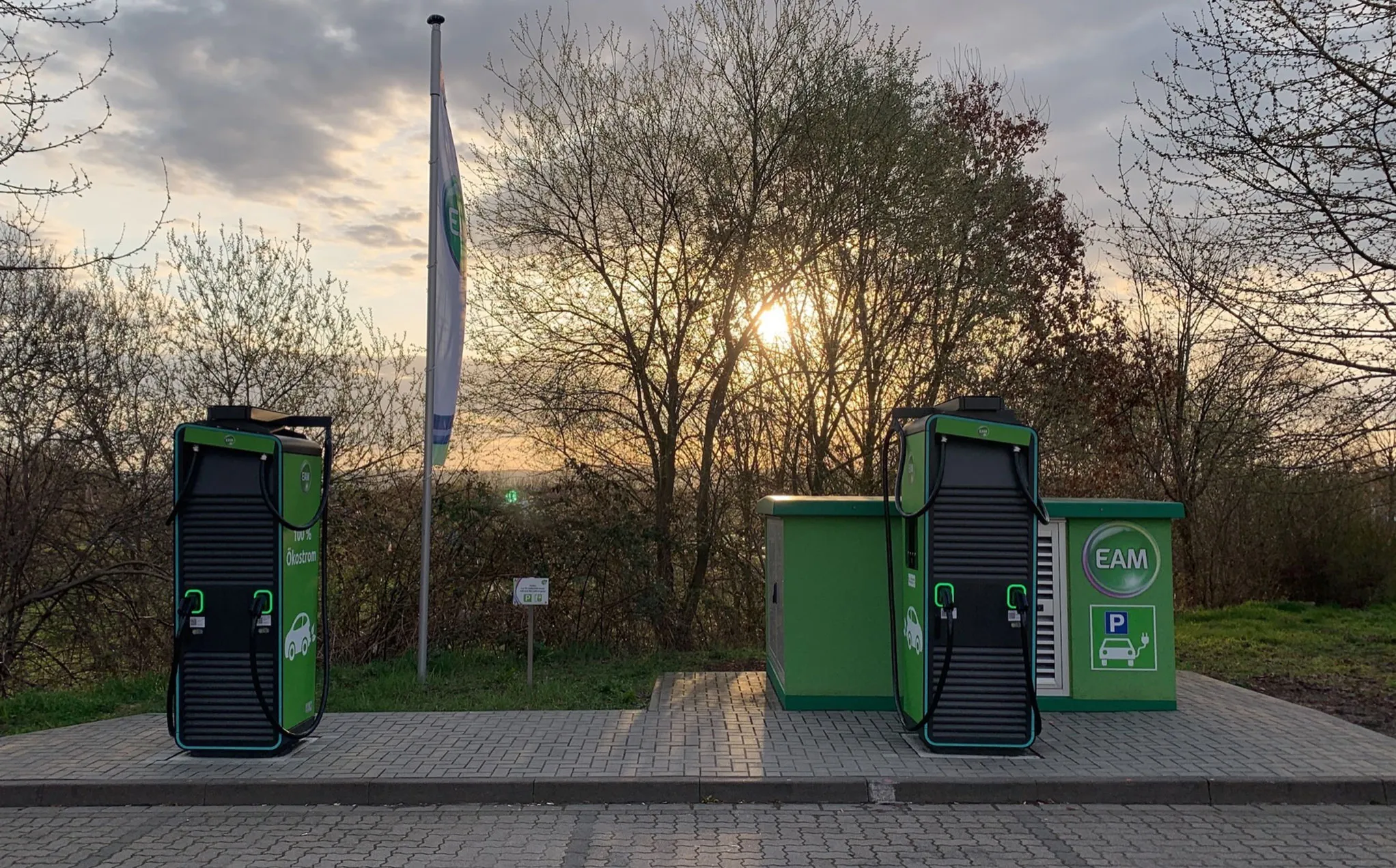 Ladesäulen für Elektroautos an einer Station, umgeben von Bäumen bei Sonnenuntergang.