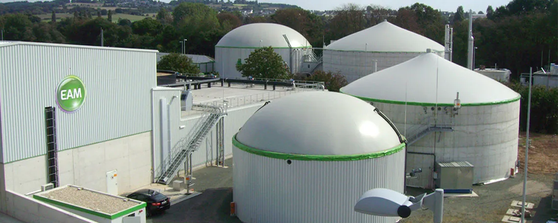 Bild einer Biogasanlage mit mehreren runden Fermentern und einem Gebäudeteil im Vordergrund, umgeben von Bäumen.