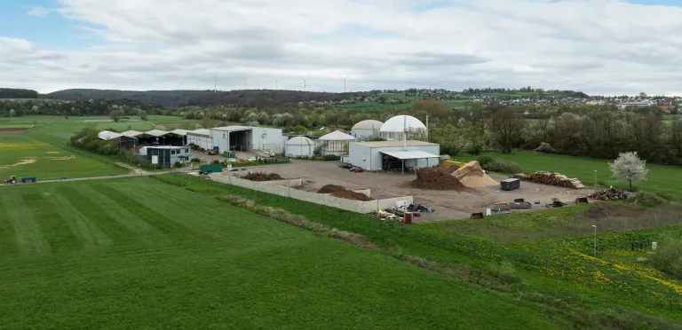 Betriebsgelände einer Biogasanlage mit mehreren Gebäuden und Freiflächen für Lagerung, umgeben von grünen Feldern und Hügeln.