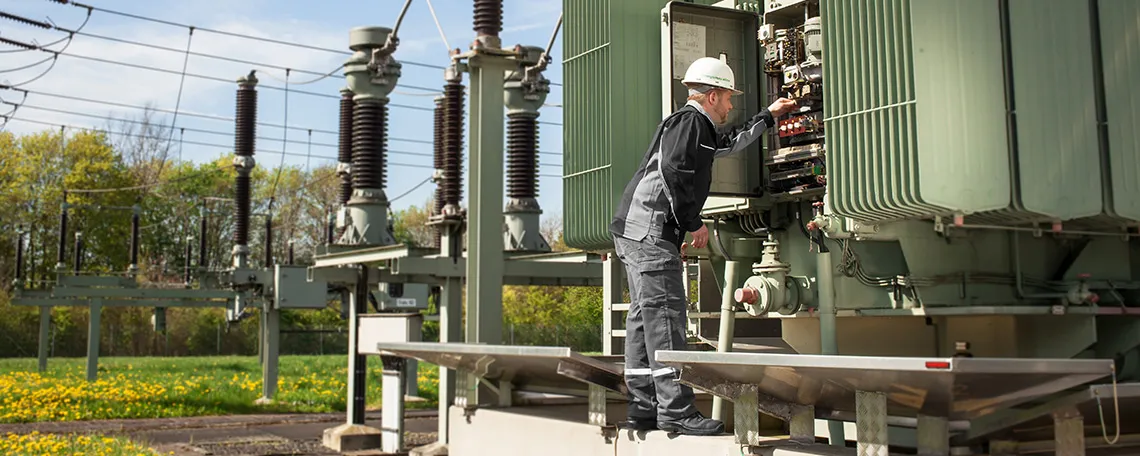 Mitarbeiter in Schutzkleidung wartet an elektrischem Transformator im Freien, umgeben von grünem Gras und Bäumen.