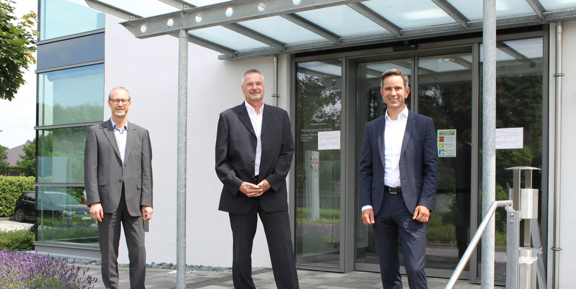 Drei Männer in Anzügen stehen vor einem modernen Gebäude mit Glasfront und lächeln..