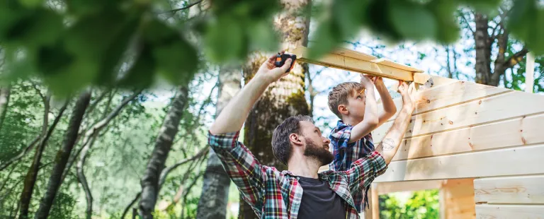 Ein Erwachsener und ein Kind befestigen gemeinsam Holzbretter an einem kleinen Holzhaus im Garten.