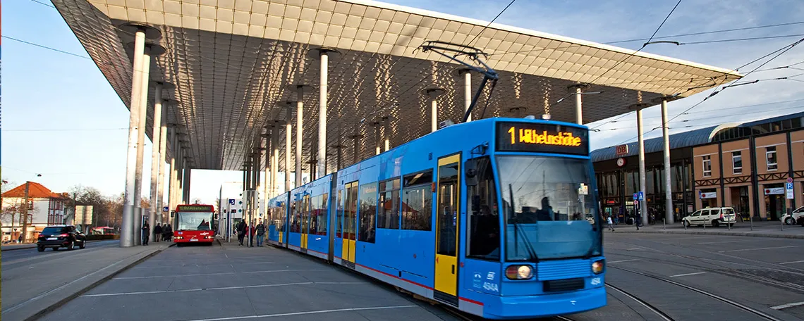 Blaue Straßenbahn und roter Bus halten an einer modernen Haltestelle mit flachem Dach und vielen Fahrgästen.