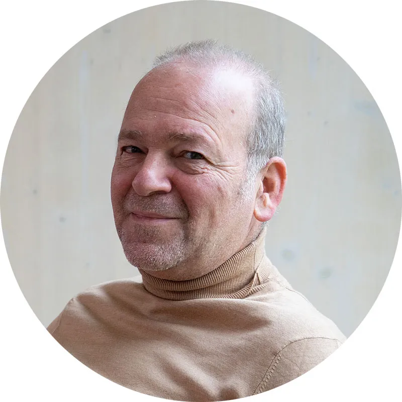 Porträt eines lächelnden Mannes mit kurzem grauen Haar, der einen beige Pullover trägt, vor hellem Hintergrund.