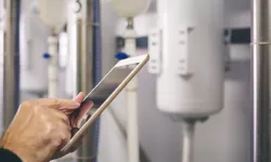 Eine Hand hält ein Smartphone vor Rohrleitungen und einer weißen Maschine in einem technischen Umfeld.