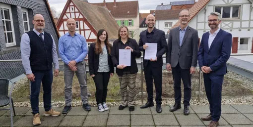 Acht Personen stehen im Freien vor Fachwerkhäusern; zwei Personen halten unterzeichnete Dokumente in die Kamera. Die Gruppe steht auf einer gepflasterten Fläche, im Hintergrund sind Gebäude und Dächer zu sehen.