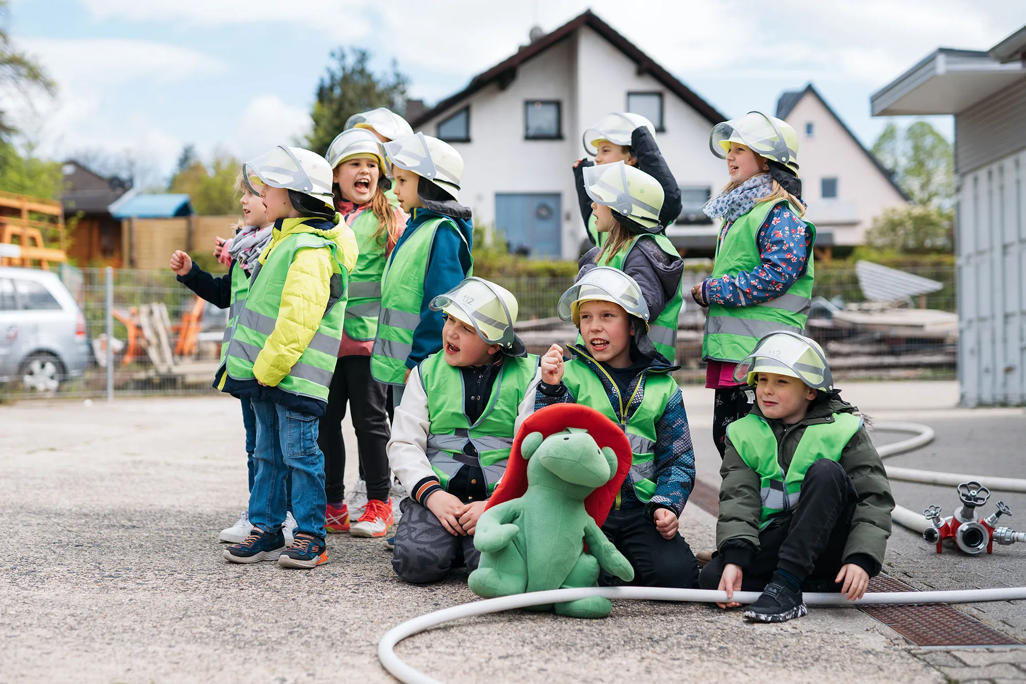 Gruppe von Kindern in Sicherheitswesten und Helmen, lachend und posierend, mit einem grünen Plüschtier auf dem Boden.