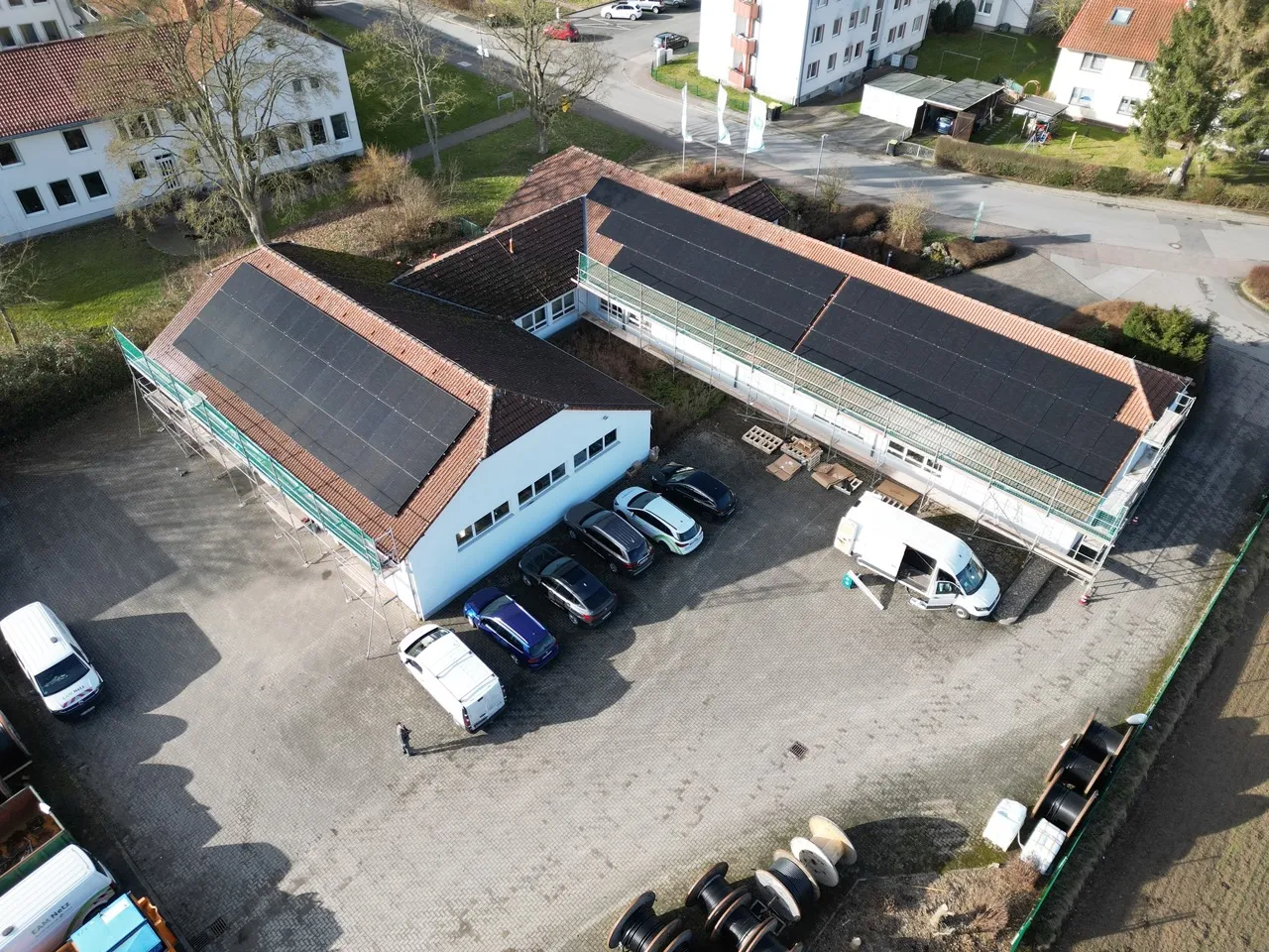 Luftaufnahme eines Gebäudes mit Solarpanelen auf dem Dach, umgeben von Parkplätzen und Bäumen.