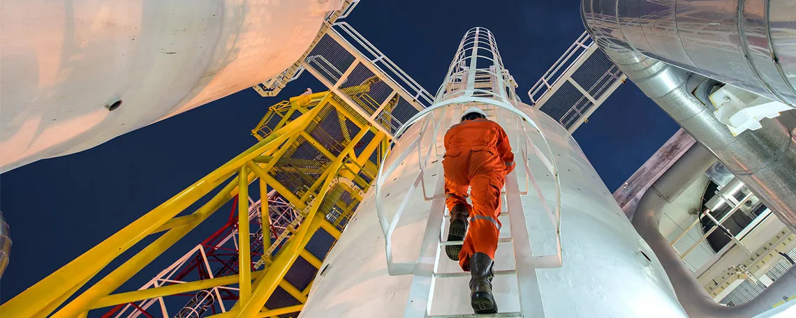 Ein Arbeiter in orangefarbener Arbeitskleidung klettert eine Leiter an einem industriellen Behälter hoch.