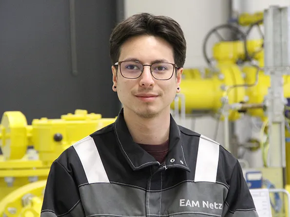 Junger Mann mit Brille, in Arbeitskleidung, steht vor einer technischen Anlage in einem industriellen Umfeld.