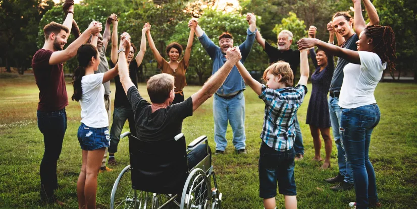 Eine Gruppe von Menschen unterschiedlichen Alters und mit unterschiedlichen Hautfarben steht im Freien auf einer Wiese in einem Kreis und hält sich an den Händen; eine Person sitzt im Rollstuhl. Im Hintergrund sind Bäume und Grünflächen bei Tageslicht zu sehen.