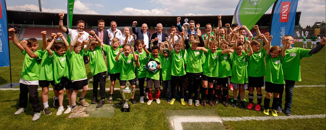 Gruppe jubelnder Kinder in grünen Trikots, die einen Pokal auf einem Fussballfeld halten.