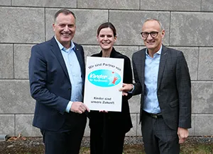 Der EAM-Geschäftsführer Olaf Kieser, die Leiterin des Bereichs Sponsoring der EAM und der Chef der HNA stehen vor einer grauen Steinwand und halten gemeinsam ein Schild. Auf dem Schild steht: ‚Wir sind Partner von Kinder für Nordhessen – Kinder sind unsere Zukunft‘, begleitet von einem Logo mit einer stilisierten Figur und blauen sowie roten Akzenten.
