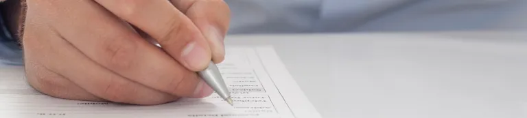 Nahaufnahme einer Hand, die mit einem Stift auf einem Formular mit vorgedruckten Zeilen und Feldern schreibt.