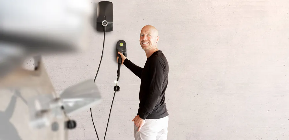 Mann lächelt, während er eine Elektroauto-Ladestation an einer Wand bedient. Moderne, minimalistische Umgebung.