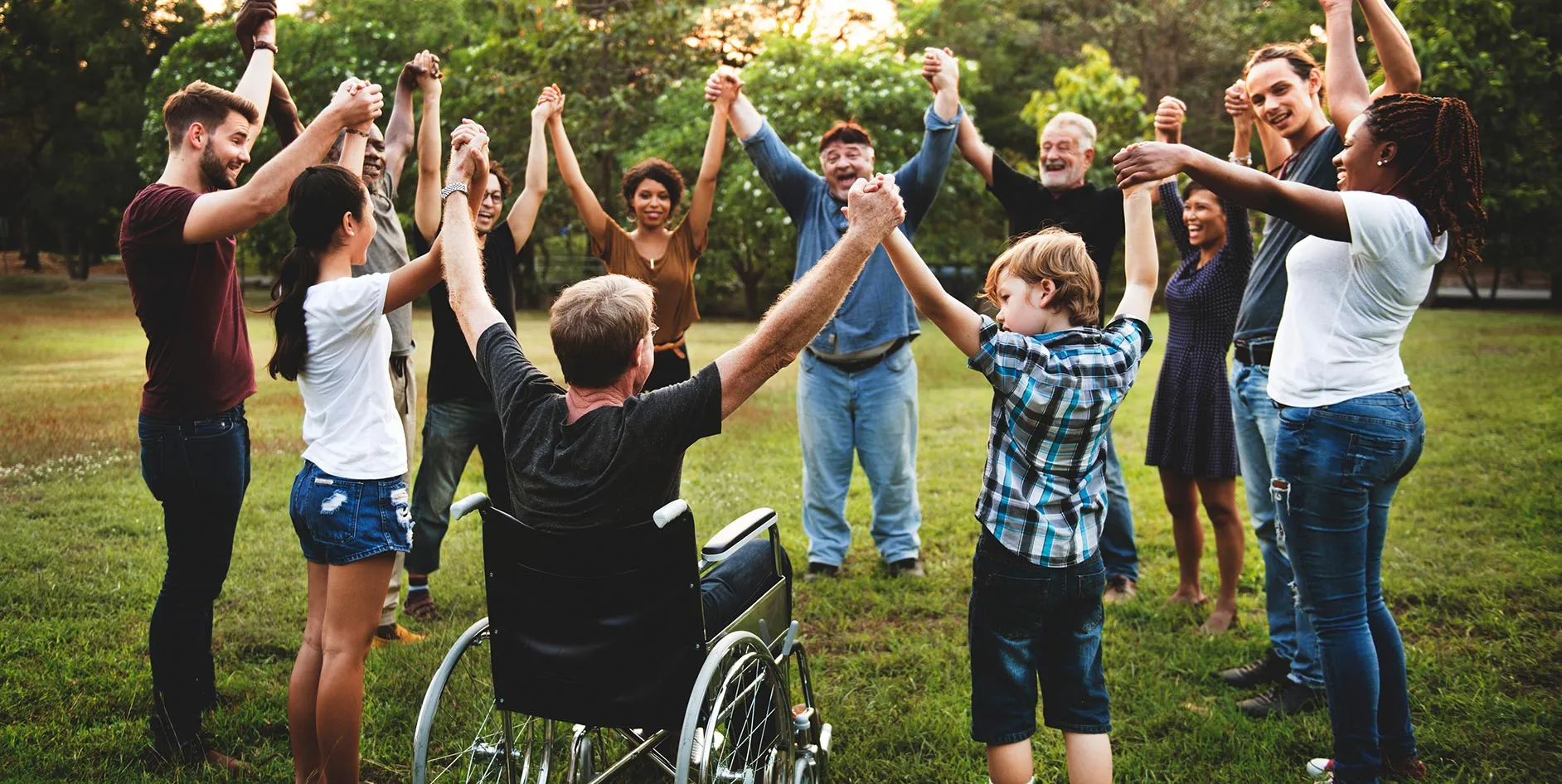 Eine Gruppe von Menschen unterschiedlichen Alters und mit unterschiedlichen Hautfarben steht im Freien auf einer Wiese in einem Kreis und hält sich an den Händen; eine Person sitzt im Rollstuhl. Im Hintergrund sind Bäume und Grünflächen bei Tageslicht zu sehen.