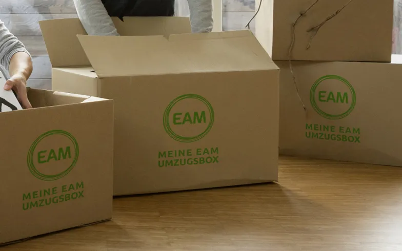 Zwei Personen packen Kartons mit dem Logo EAM und dem Text MEINE EAM UMZUGSBOX auf einem hellen Holzfußboden.