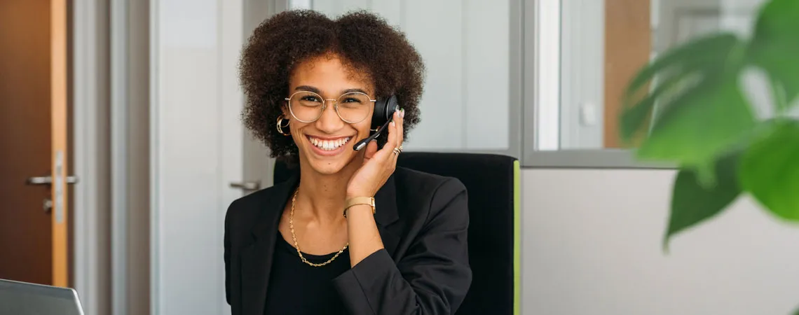 Lächelnde Person mit Headset in einem modernen Büro, sitzt vor einem Laptop und spricht am Telefon.
