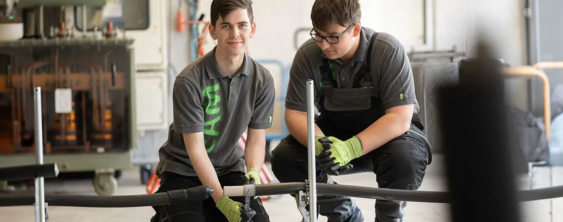 Zwei junge Männer arbeiten in einer Werkstatt an einem technischen Projekt mit einem Rohr und Werkzeug.