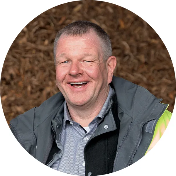Lächelnder Mann in Arbeitskleidung mit grauer Jacke vor einem Hintergrund aus Holzspänen.