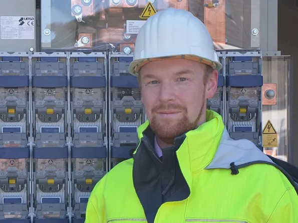 Porträt eines Mannes in gelber Sicherheitsjacke und Helm vor einem Schaltpanel mit Stromzählern.