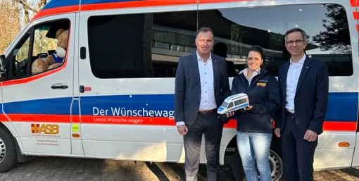 Die EAM-Geschäftsführer Olaf Kieser und Hans-Hinrich Schriever übergeben die Spende an die Projektkoordinatorin des Wünschewagens Nordhessen Larissa Groffmann vor dem Wünschewagen, an dessen Steuer ein riesiger Teddy sitzt.