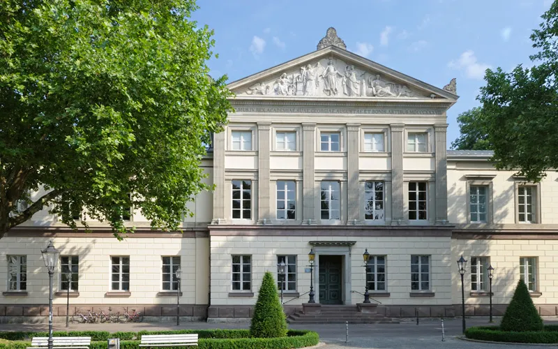 Hübsches historisches Gebäude mit klassizistischer Fassade, umgeben von Bäumen und einer gepflegten Grünfläche.