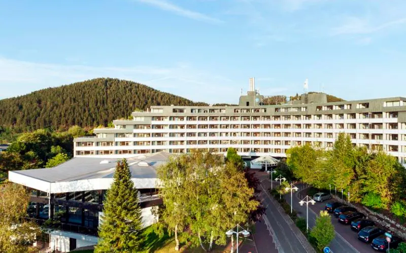 Panoramablick auf ein modernes Hotelgebäude mit Parkplätzen, umgeben von Bäumen und Hügeln.