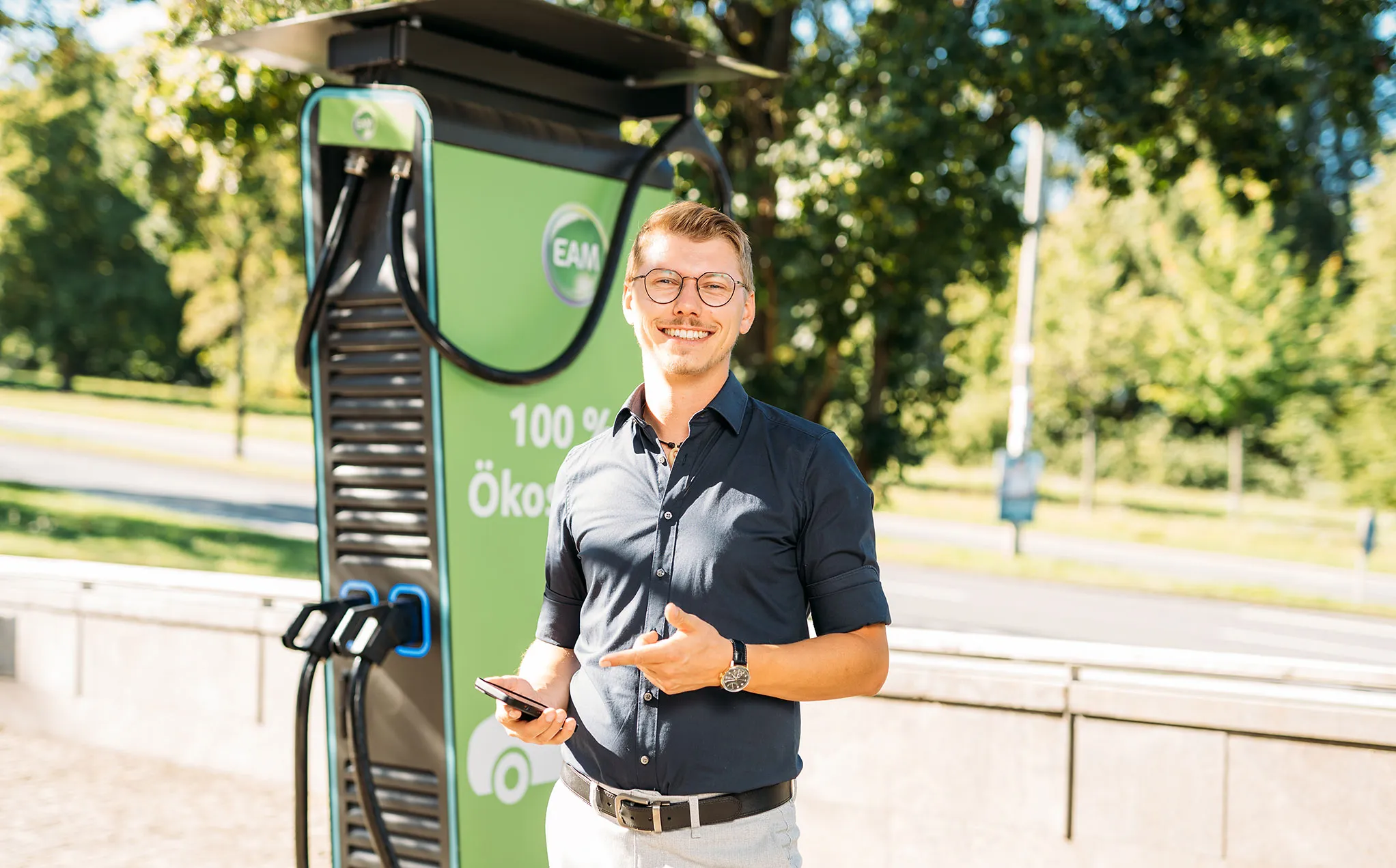 Lächelnder Mann mit Smartphone neben einer grünen Ladestation für Elektroautos im Freien.