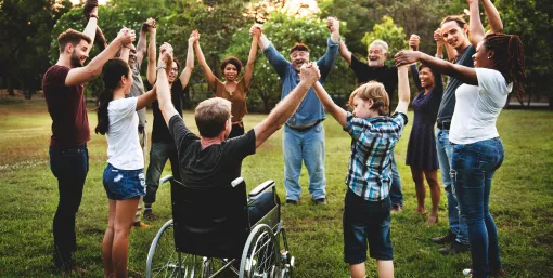 Eine Gruppe von Menschen unterschiedlichen Alters und mit unterschiedlichen Hautfarben steht im Freien auf einer Wiese in einem Kreis und hält sich an den Händen; eine Person sitzt im Rollstuhl. Im Hintergrund sind Bäume und Grünflächen bei Tageslicht zu sehen.