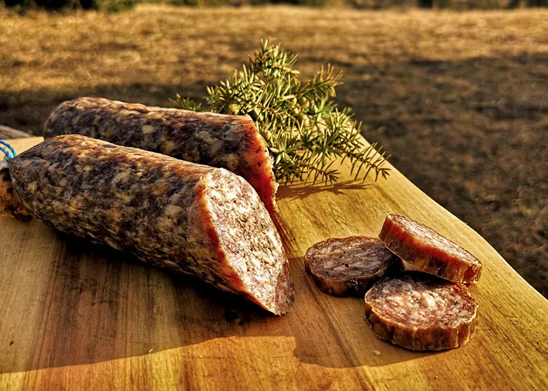 Zwei Stücke Fleischwurst auf einem Holzbrett, frisch geschnitten, umgeben von grünen Kräutern.