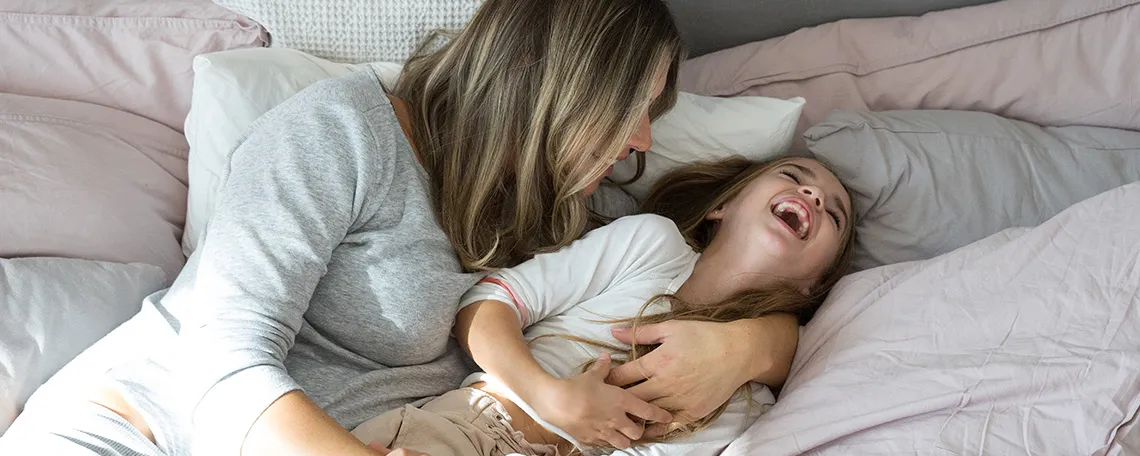 Eine Mutter und ihre Tochter lachen gemeinsam auf einem Bett, umgeben von Kissen und Bettwäsche.