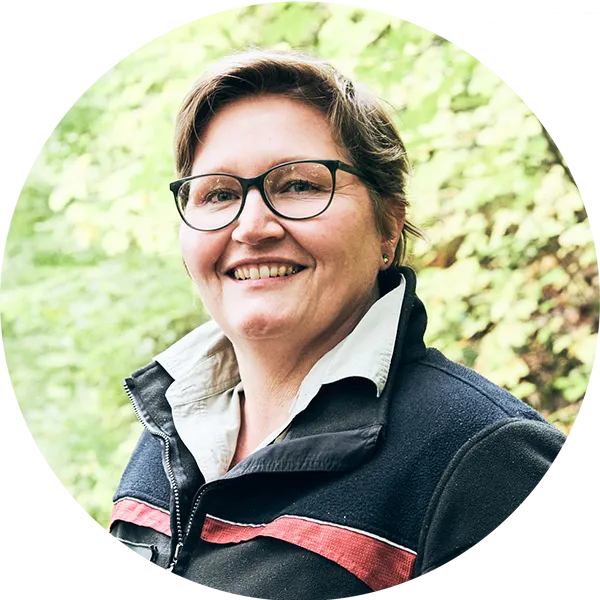 Porträt einer lächelnden Frau mit Brille und Fleecejacke, umgeben von grüner Natur.