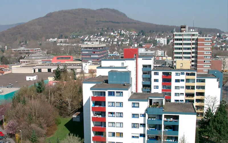 Stadtansicht mit Wohngebäuden und Hügel im Hintergrund, klare Tageslichtaufnahme.