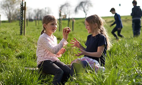 Zwei Mädchen sitzen im Gras und spielen mit einem Apfel, während andere Kinder im Hintergrund spielen.