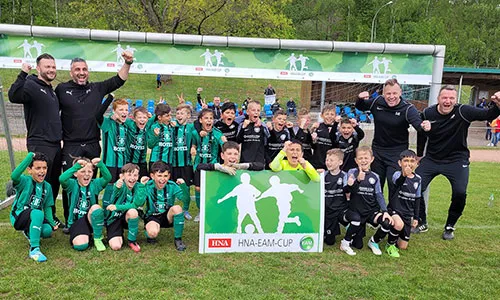 Ein Gruppenfoto von Fußballkindern und Trainern, die jubelnd vor einem Banner des HNA-EAM-Cups stehen.