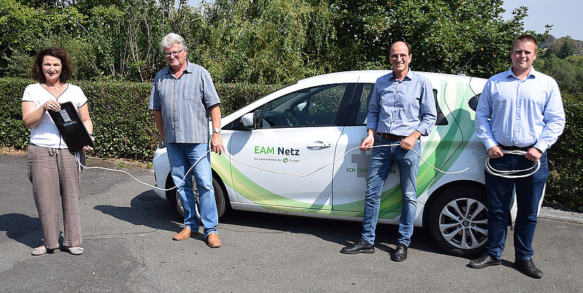 Vier Personen stehen an einem Elektroauto, während sie Ladevorrichtungen halten. Im Hintergrund grüne Bäume.