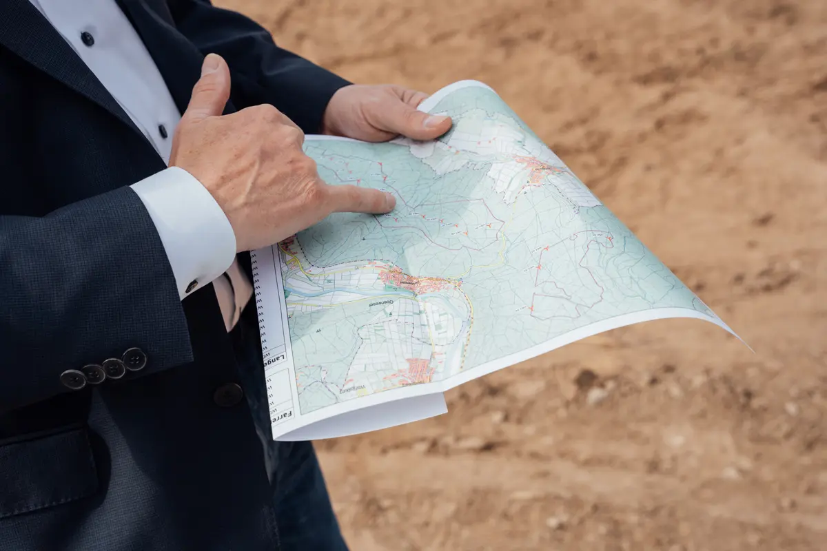 Person in Anzug hält eine Landkarte und zeigt mit dem Finger auf einen bestimmten Punkt. Hintergrund ist brauner Boden.