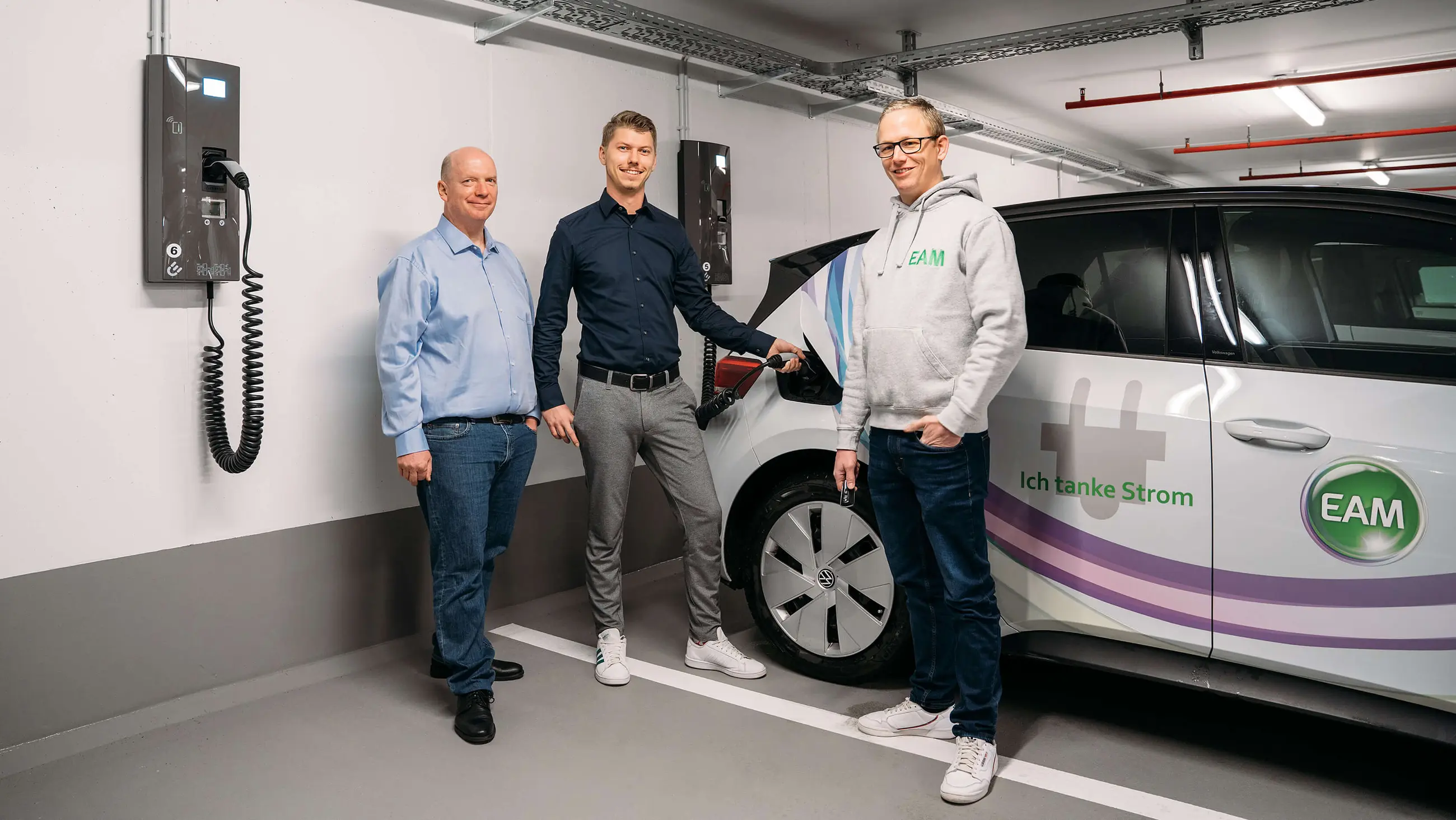 Drei Männer stehen an einer Ladestation für Elektroautos und halten ein Ladekabel in der Hand.