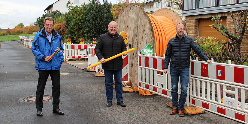 Drei Männer stehen an einer Baustelle mit Kabelrolltrommeln und Absperrungen in einer Wohngegend.