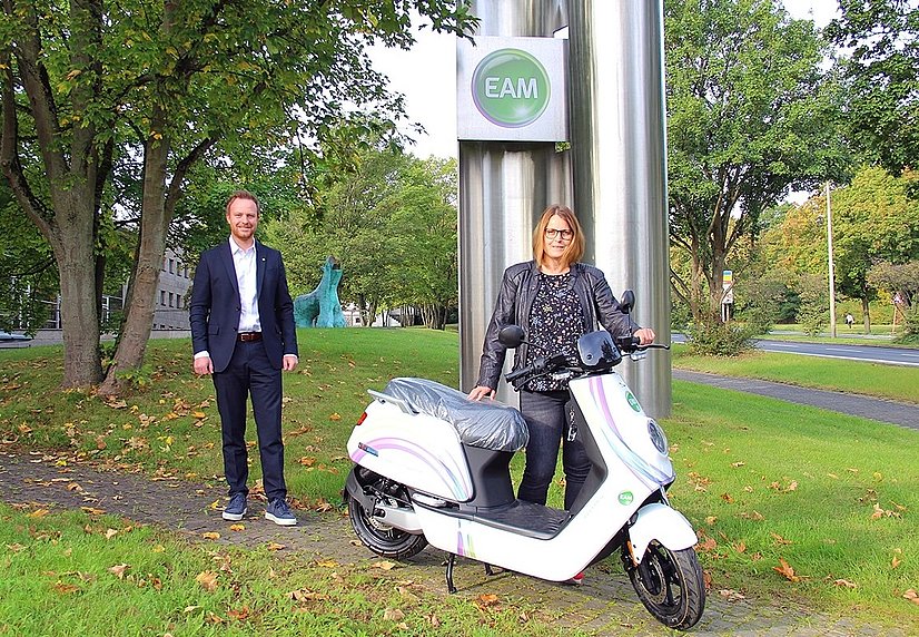 Manuela Sauer steht neben einem weißen Elektroroller, ein Mann steht lächelnd daneben, im Hintergrund ein EAM-Schild.