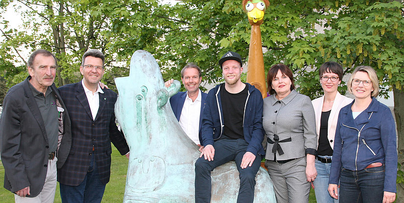 Ein Gruppe von sieben Personen, fünf Männer und zwei Frauen, posiert im Freien um eine abstrakte Skulptur.