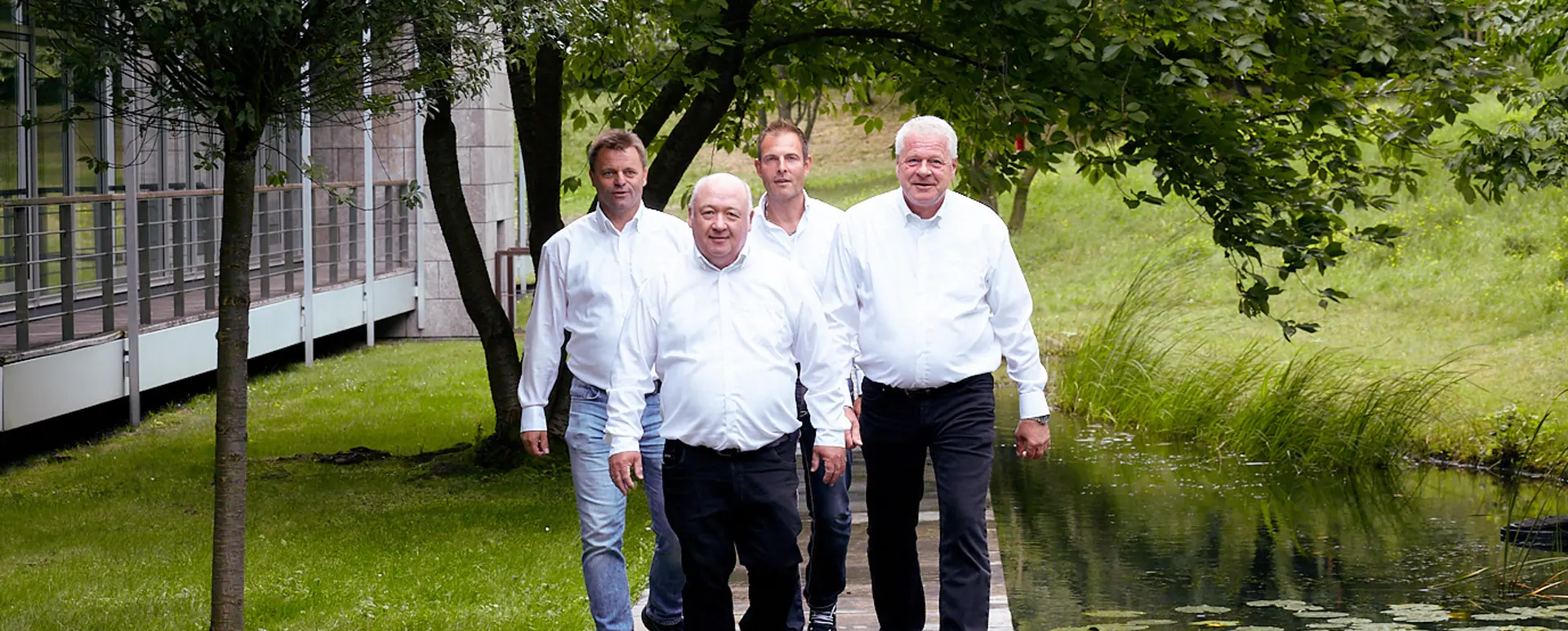 Vier Männer in weißen Hemden gehen am Wasser entlang, umgeben von Bäumen und Gras.