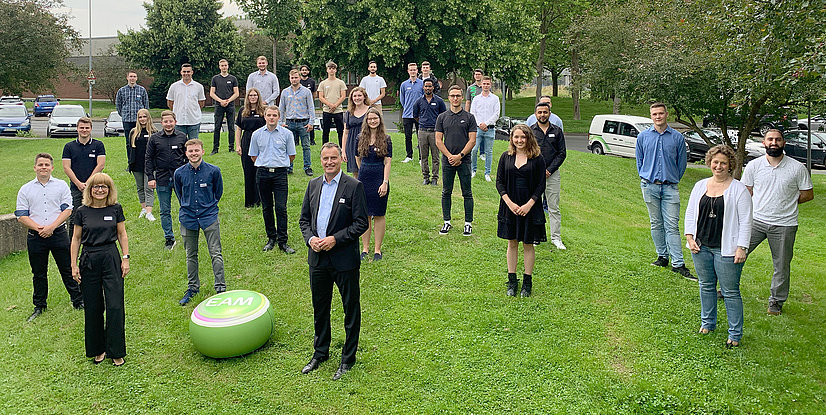 Großes Gruppenfoto der ausgelernten Auszubildenden auf einer Wiese, einige tragen Namensschilder, EAM-Logo auf einem grünen Ball im Vordergrund.