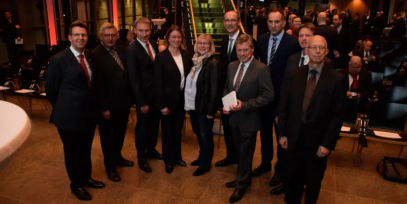 Eine Gruppe von zehn Personen posiert in einem modernen Veranstaltungsraum. Es gibt Stühle und Beleuchtung im Hintergrund.