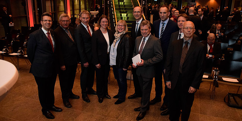 Eine Gruppe von zehn Personen posiert in einem modernen Veranstaltungsraum. Es gibt Stühle und Beleuchtung im Hintergrund.