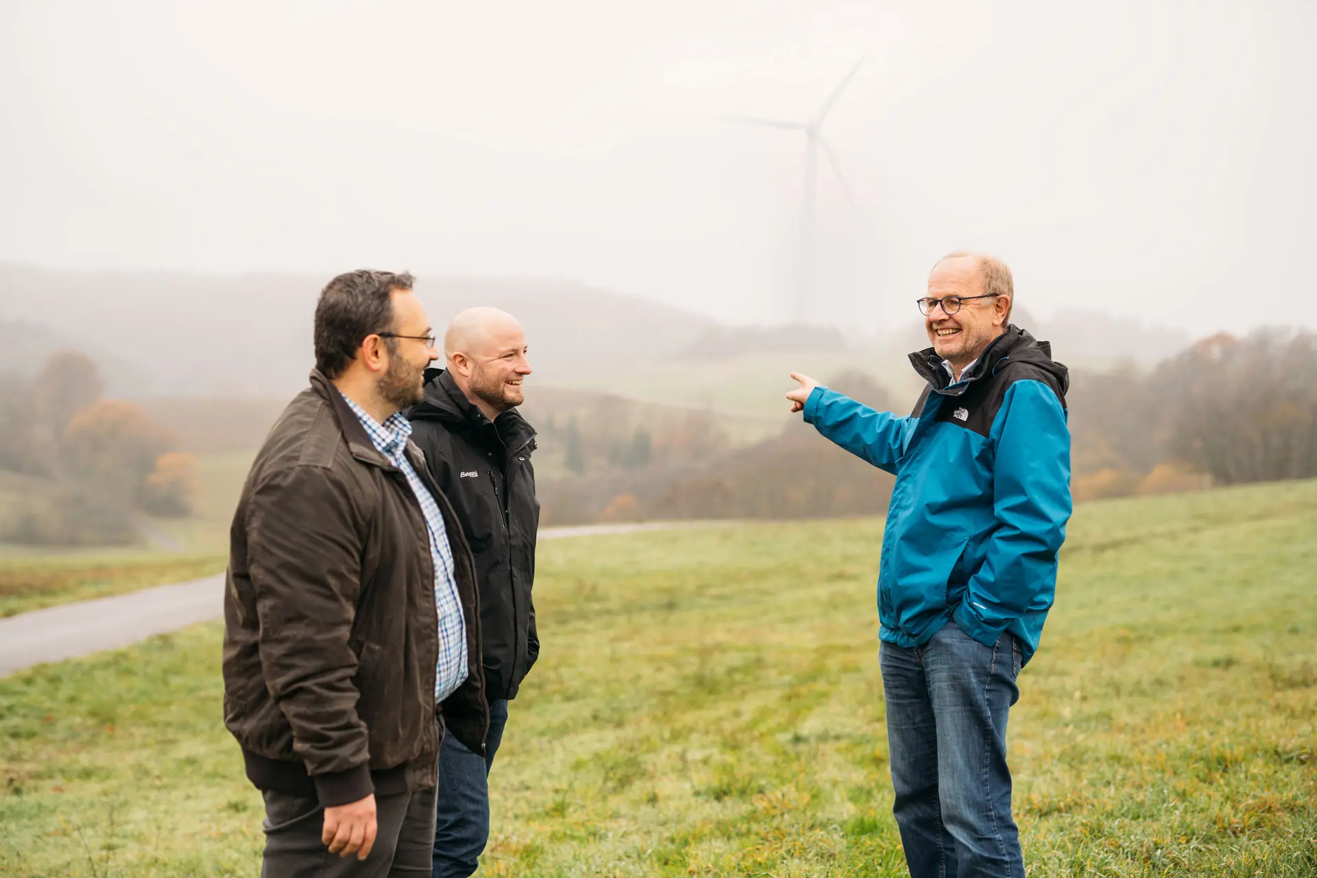 Drei Männer stehen im Freien, auf einem sanften Hügel mit Windrädern im Hintergrund, in einer nebligen Landschaft.