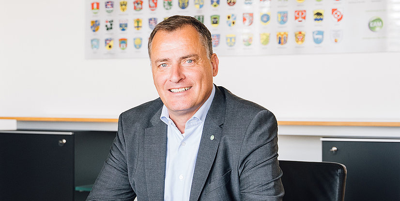 Der Geschäftsführer der EAM Olaf Kieser sitzt lächelnd vor einer Wand mit mehreren Wappen.