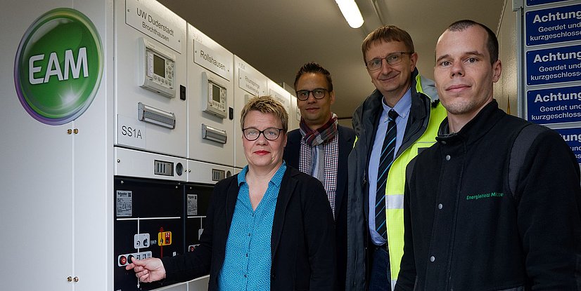 Vier Personen stehen vor einem elektrischen Verteiler mit dem EAM-Logo. Eine Person betätigt einen Schalter.
